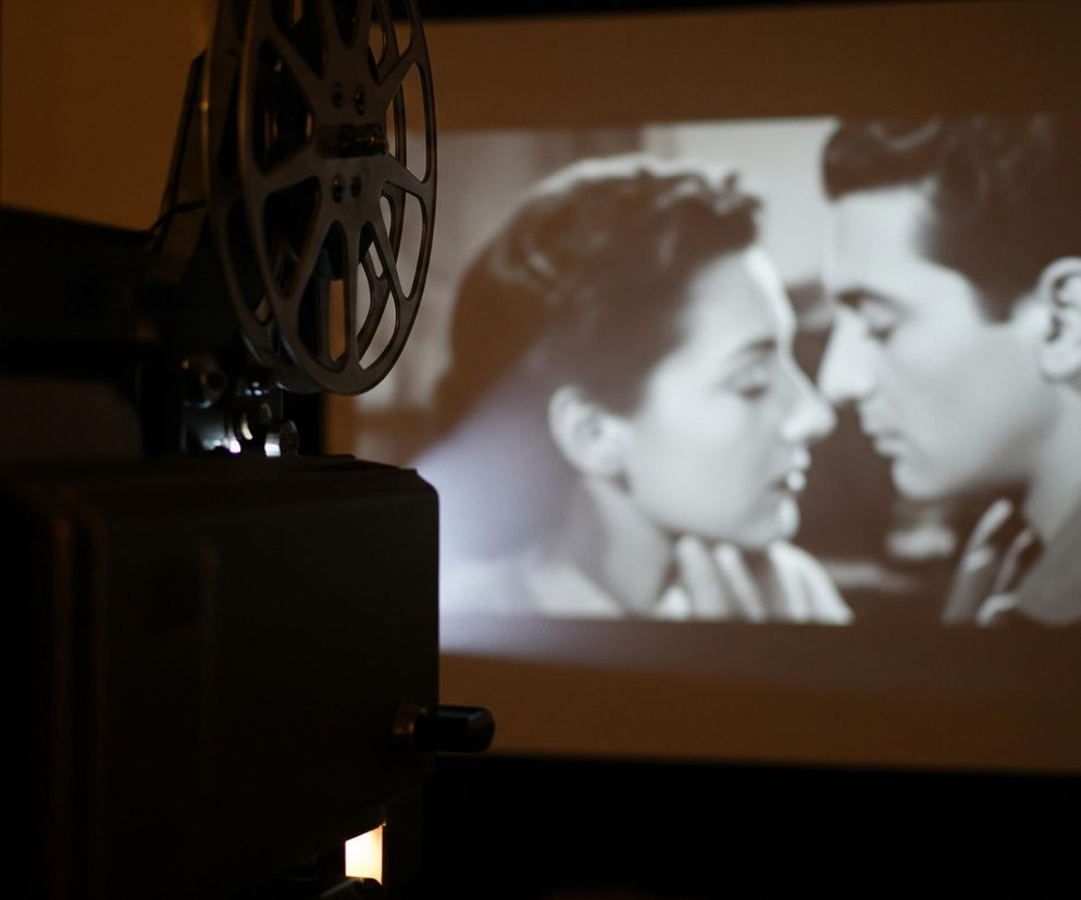 Czarny projektor filmowy, widoczny częściowo po lewej stronie kadru, rzuca światło na jasną powierzchnię, na której wyświetla się czarno-biała scena z filmu. Na ekranie widać zbliżenie dwóch osób, mężczyzny i kobiety, spoglądających na siebie z bliskiej odległości. Kobieta delikatnie dotyka podbródka mężczyzny. Dominują ciemne brązy i czerń w tle, kontrastujące z jasnym, wyświetlanym obrazem.