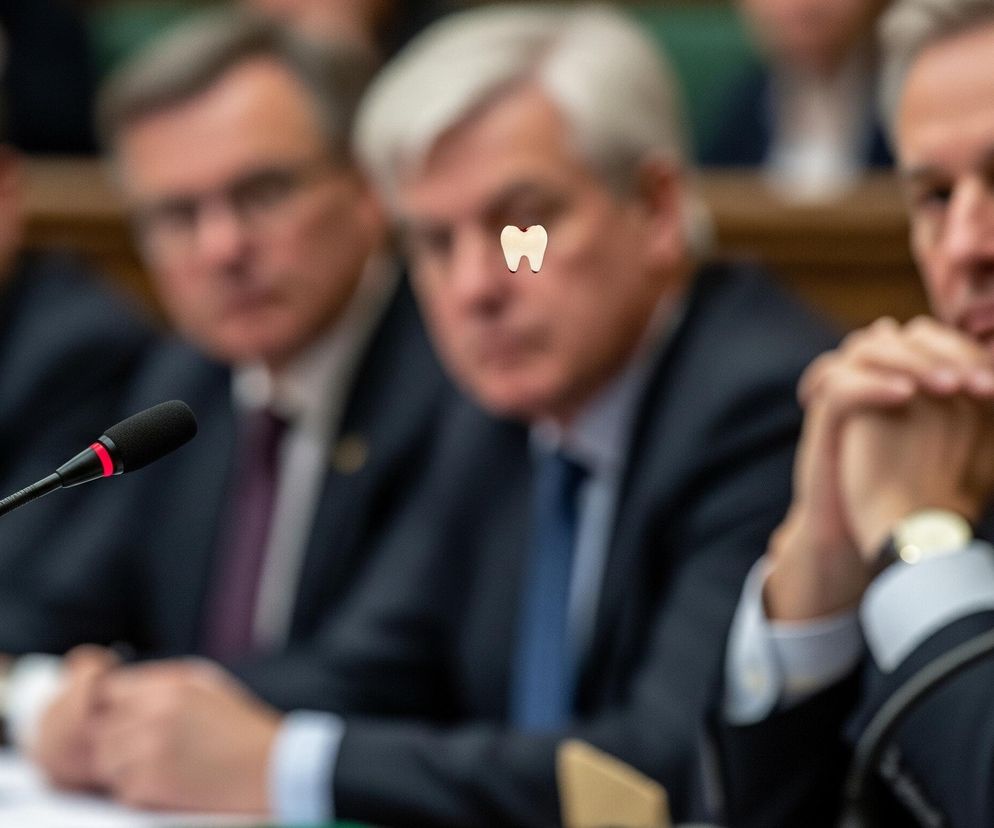 Na pierwszym planie, z lewej strony, widoczny jest czarny mikrofon na giętkiej szyjce, z czerwoną opaską na zakończeniu. Za mikrofonem, w tle, znajduje się czterech mężczyzn w ciemnych garniturach i krawatach, siedzących obok siebie przy stole. Trzech mężczyzn z lewej strony ma zamazane twarze, a mężczyzna po prawej stronie jest częściowo widoczny z zaciśniętymi rękami. Nad głową drugiego mężczyzny od prawej, który ma siwe włosy, unosi się mały, biały obiekt w kształcie zęba.