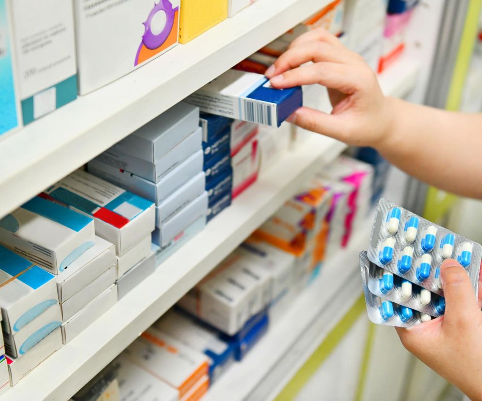 Ręka farmaceuty lub osoby obsługującej aptekę sięga po opakowanie leku z półki, na tle regałów z różnymi medykamentami. W drugiej dłoni trzyma blistry z niebiesko-białymi kapsułkami, co symbolizuje proces wydawania lub sprawdzania leków w kontekście wycofania popularnego leku na tarczycę, o czym przeczytasz na Poradnik Zdrowie.