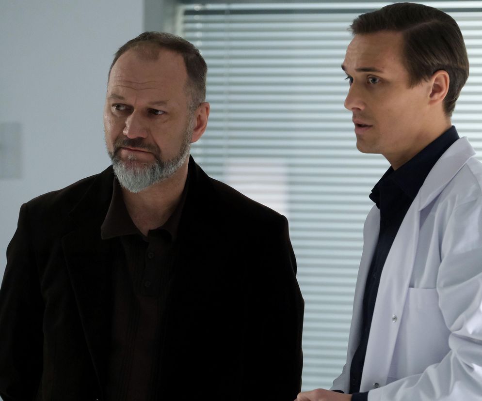 Mirosław Wójcik (Jakub Kornacki) i Olek Chodakowski (Maurycy Popiel) w scenie z serialu „M jak miłość”. Olek w białym kitlu, a Wójcik w ciemnym płaszczu stoją obok siebie w szpitalu. Więcej o dramatycznych losach bohaterów przeczytasz na Super Seriale.