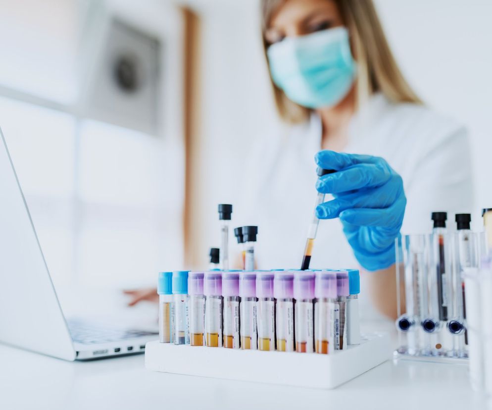 Osoba w masce ochronnej i niebieskich rękawiczkach laboratoryjnych trzyma probówkę z próbką krwi. Przed nią na stole znajduje się stojak z licznymi probówkami, a obok otwarty laptop, co sugeruje badania naukowe. Więcej o bisfenolu A i jego wpływie na zdrowie przeczytasz na Poradnik Zdrowie.