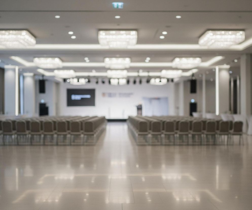 W przestronnej, jasno oświetlonej sali konferencyjnej, z parkietem odbijającym światło, ustawione są rzędy krzeseł z ciemnymi obiciami, tworząc dwie sekcje widowni. Z przodu znajduje się scena z ekranem, na którym widoczne są niewyraźne napisy i grafiki. Sufit zdobią liczne, kwadratowe żyrandole oraz wpuszczone w jego strukturę listwy LED, a po bokach, na całej długości pomieszczenia, rozmieszczone są wysokie, białe słupy z pionowymi pasami oświetlenia. Przy ścianach widoczne są również białe, wysokie stoliki koktajlowe, oraz zasłonięte ciemnymi zasłonami okna.