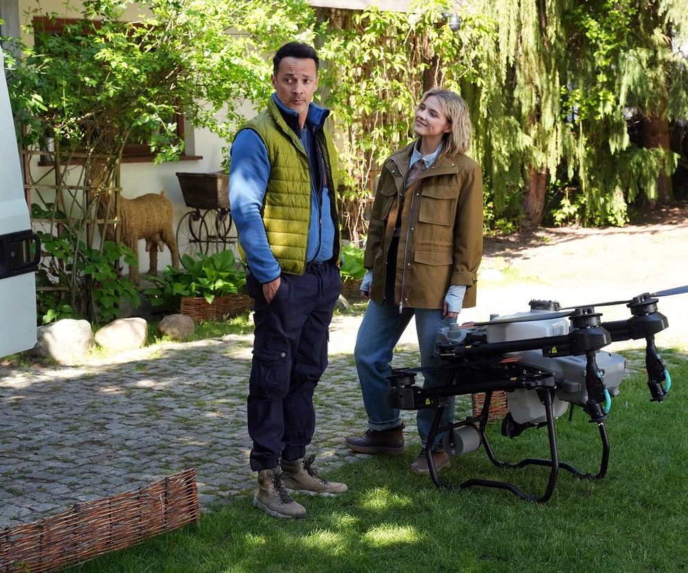 Mariusz (Rafał Cieszyński) i Janka (Oliwia Drożdżyk) z serialu Barwy szczęścia rozmawiają na podwórku obok drona rolniczego. Janka w brązowej kurtce i jeansach uśmiecha się zalotnie do Mariusza w niebieskiej bluzie i zielonej kamizelce. Więcej o ich relacji przeczytasz na Super Seriale.