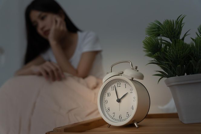 Biały budzik na stoliku nocnym, wskazujący godzinę drugą, przed którym siedzi smutna, młoda kobieta z bezsennymi oczami, z niewyraźną miną w tle, w półmroku, symbolizujący problemy ze snem i ryzyko demencji, o których możesz przeczytać na portalu Poradnik Zdrowie.