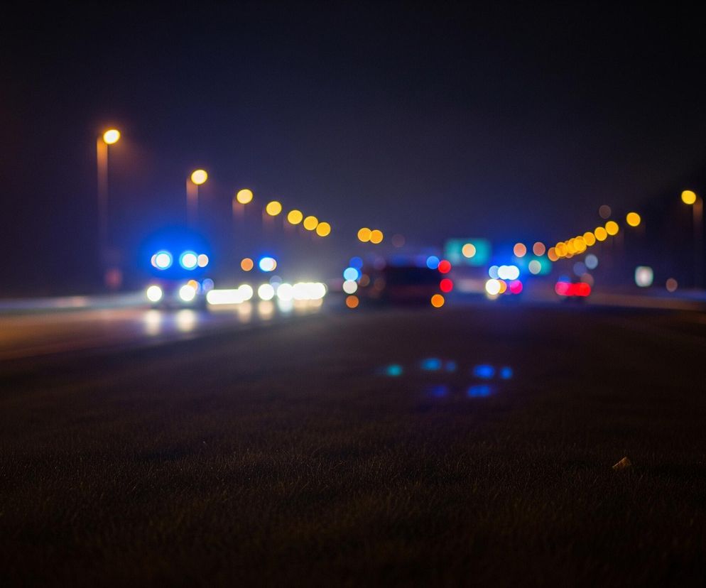 Rozmyte, nocne ujęcie autostrady, z ciemnym asfaltem na pierwszym planie pokrywającym dolną połowę obrazu. W tle widać rzędy jasnych, ciepłych pomarańczowych lamp ulicznych i świateł samochodów, głównie białych, niebieskich i czerwonych, tworzących świetliste smugi i kręgi w wyniku efektu bokeh. Po lewej stronie widoczne są wydłużone smugi jasnych świateł, a po prawej delikatne, rozmyte kształty znaków drogowych z zielonymi i białymi elementami.