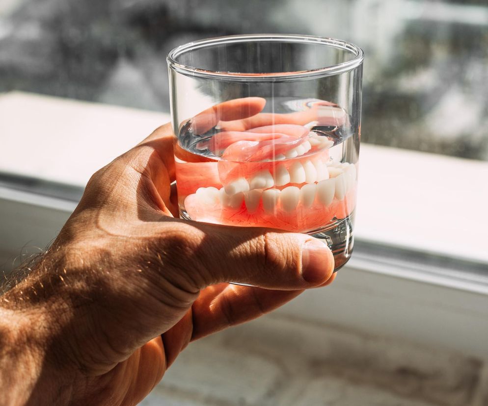 Męska dłoń trzyma szklankę z wodą, w której zanurzona jest różowa proteza zębowa, symbolizująca problem bezzębia w Polsce, na tle jasnego okna. Więcej o zagrożeniach dla zdrowia zębów przeczytasz na Poradnik Zdrowie.