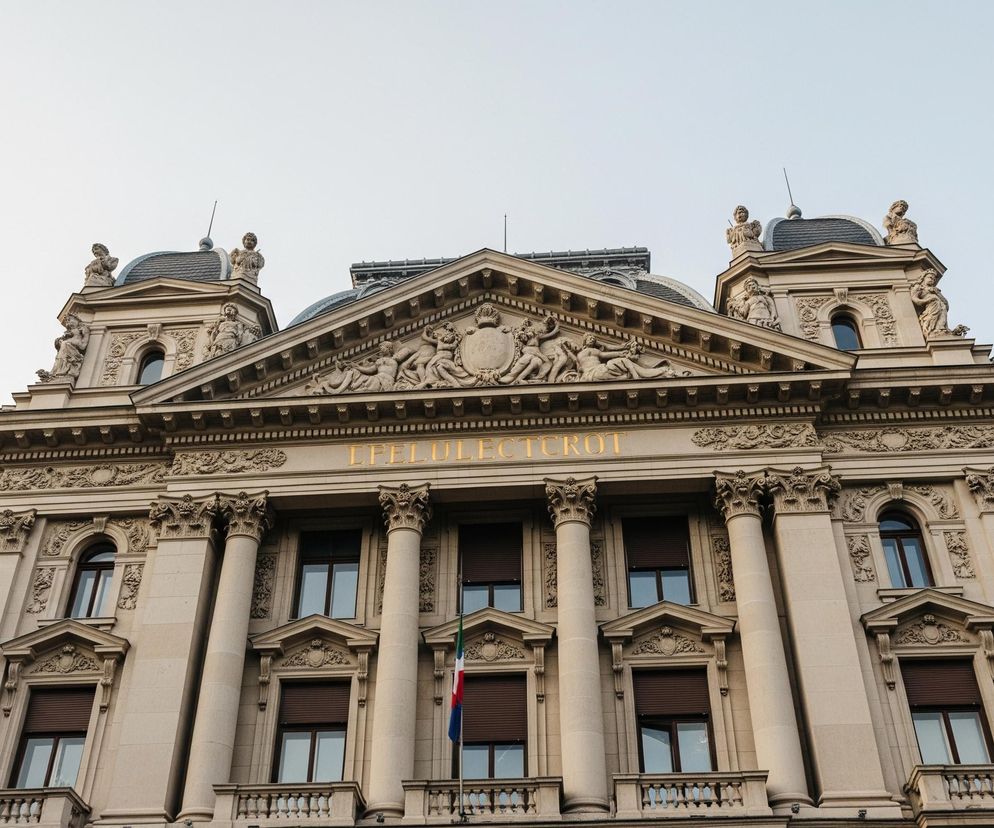 Centralnie umieszczony na zdjęciu jest duży, ozdobny beżowy budynek, którego fasada pokryta jest bogatymi detalami architektonicznymi. Górna część budynku wieńczy szczyt z płaskorzeźbami przedstawiającymi postacie ludzkie i symboliczne elementy, a poniżej widnieje złoty napis LIFEI DELECTROTUI. Fasada posiada rząd kolumn korynckich, a między nimi liczne okna z brązowymi okiennicami, z których niektóre są zamknięte. Przed jednym z centralnych okien powiewa flaga w kolorach zielonym, białym i czerwonym. Budynek posiada także ozdobne balkony z balustradami, a na dachu widoczne są kopuły z rzeźbami i szpiczaste iglice, wszystko to na tle jasnego, czystego nieba.