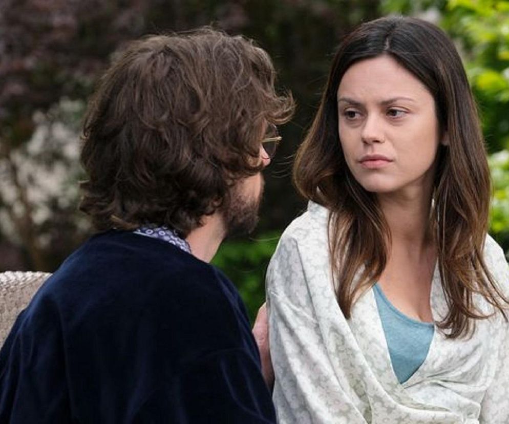 Paulina Lasota jako Kasia z M jak miłość, z poważną miną, w jasnym wzorzystym szlafroku i błękitnym topie, rozmawia z mężczyzną w ciemnym swetrze i okularach. Scena ukazuje trudne relacje, o których przeczytasz na portalu Super Seriale.