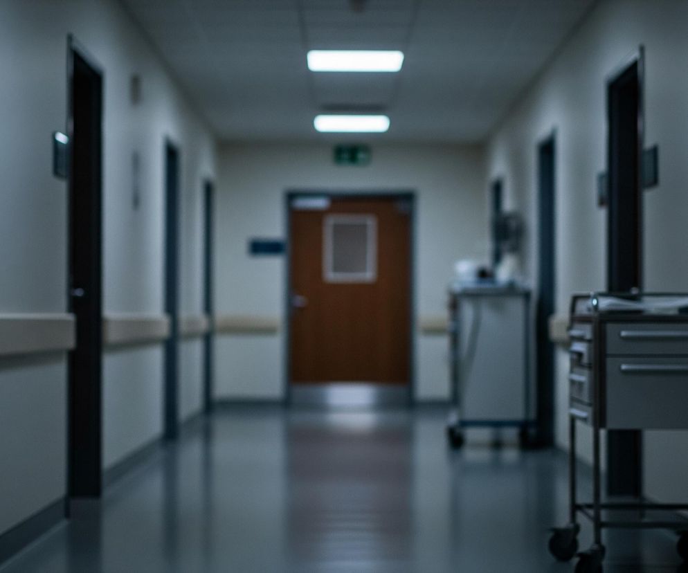 Długi korytarz szpitalny jest oświetlony od góry kilkoma prostokątnymi lampami. Po obu stronach widoczne są szare ściany z białymi, poziomymi poręczami oraz rzędy drzwi w ciemnych futrynach. Na końcu korytarza znajduje się brązowe drzwi z małym, kwadratowym okienkiem. Po prawej stronie, na pierwszym planie, stoi metalowy wózek medyczny z dwoma szufladami i blatem, na którym leżą dokumenty. Całe ujęcie jest nieostre, tworząc efekt rozmycia.