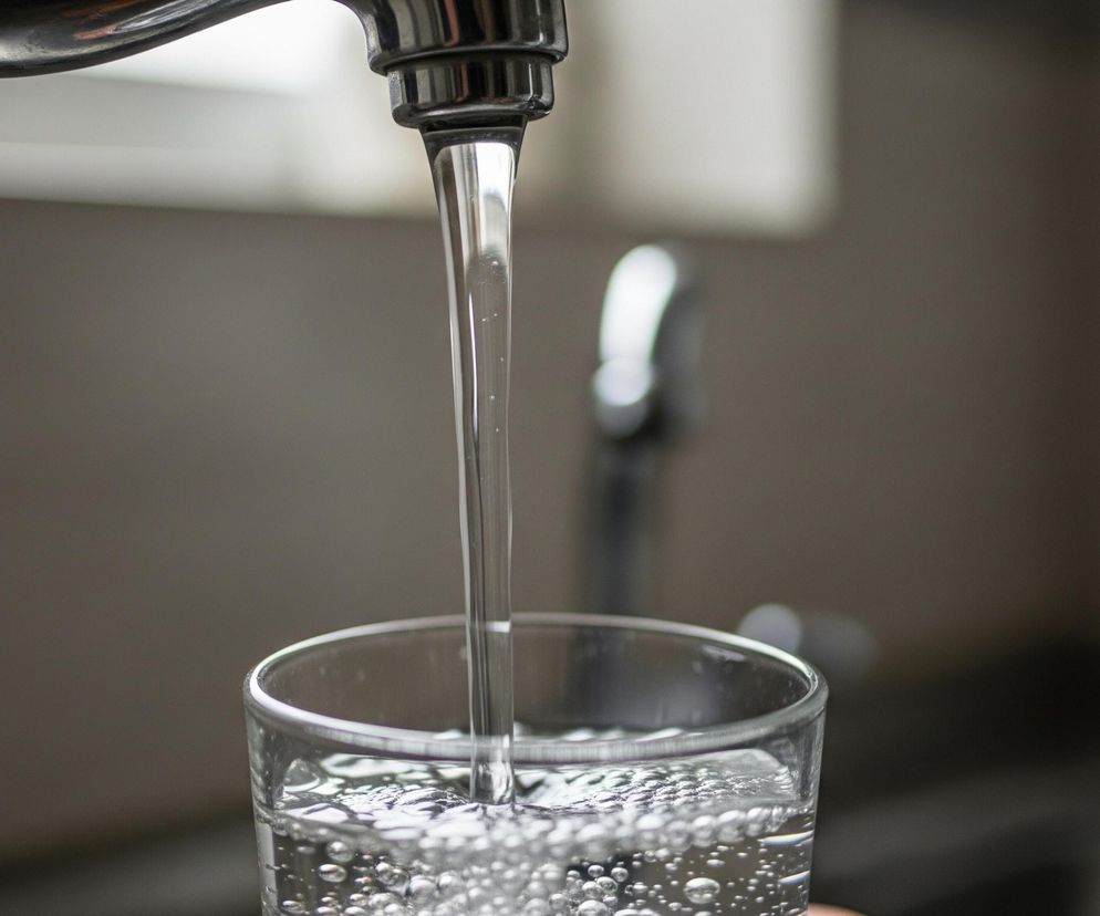 Chromowany kran, umieszczony w górnej części kadru, wylewa strumień czystej wody wprost do szklanki. Woda w szklance jest już częściowo nalana, tworząc liczne bąbelki na powierzchni i wewnątrz naczynia. Tło za kranem i szklanką jest rozmyte, utrzymane w odcieniach szarości i brązu, z jaśniejszym obszarem u góry, sugerującym okno lub inną źródło światła.