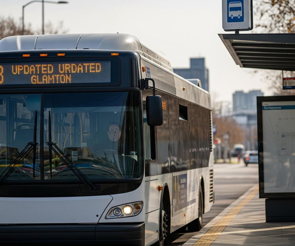 Autobus miejski w kolorze białym i czarnym, z numerem linii 103 i tekstem UPDATED GLAMTON wyświetlanym na pomarańczowym ekranie LED, znajduje się na ulicy. Widoczne są jego włączone przednie światła i numer 36 na lewej stronie zderzaka. Autobus jest zaparkowany obok przystanku z ciemną wiatą i niebieską tablicą z ikoną autobusu, a w tle widać drzewa bez liści i budynki.
