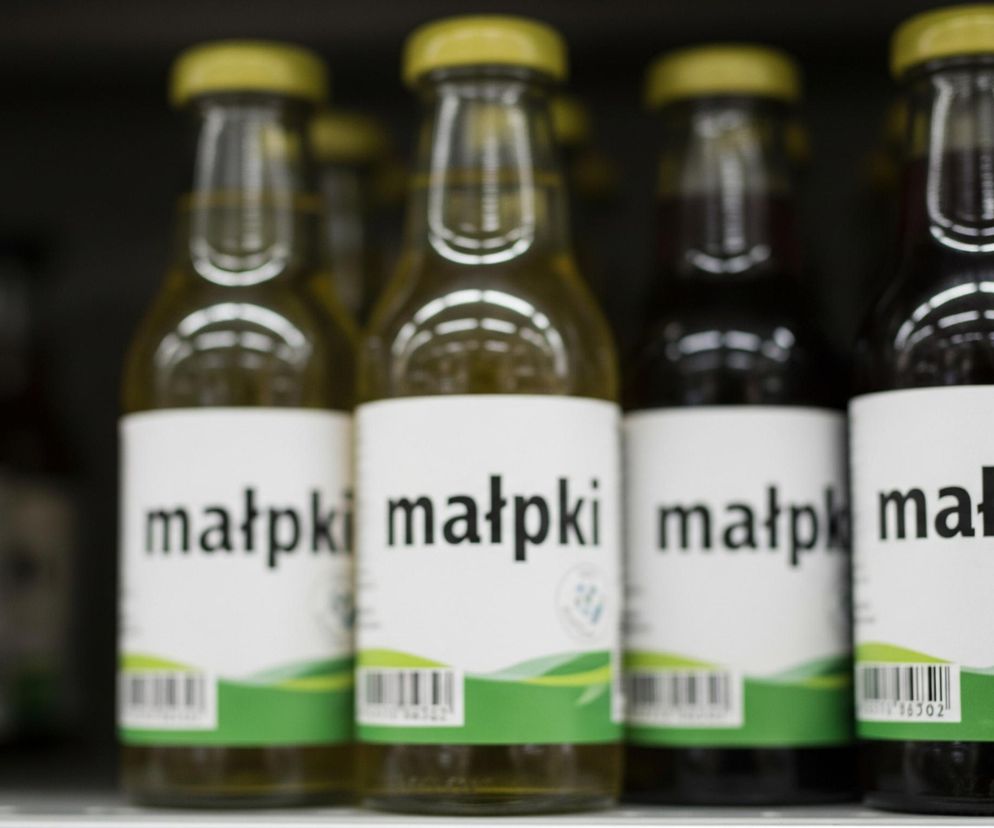 Rząd szklanych butelek z zielonymi zakrętkami stoi na półce. Na każdej butelce znajduje się biała etykieta z napisem „małpki” dużą, czarną czcionką. Poniżej napisu widoczny jest zielony falisty wzór oraz kod kreskowy. Zawartość butelek jest w różnych odcieniach, od jasnożółtego do ciemnobrązowego. Tło jest ciemne i niewyraźne, z zarysem kolejnych butelek w oddali.