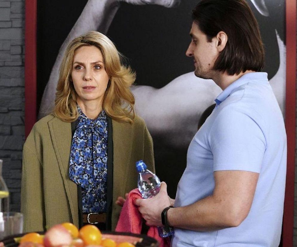 Malwina (Joanna Gleń) i Herman (Maciej Raniszewski) rozmawiają w fitness klubie. Malwina, ubrana w kwiecistą bluzkę i oliwkową marynarkę, patrzy z zaniepokojeniem. Herman, w jasnoniebieskiej koszulce, trzyma butelkę wody i ręcznik, opowiada o swoich planach, które wzbudzają jej obawy. Więcej informacji o serialu znajdziesz na stronie Super Seriale.