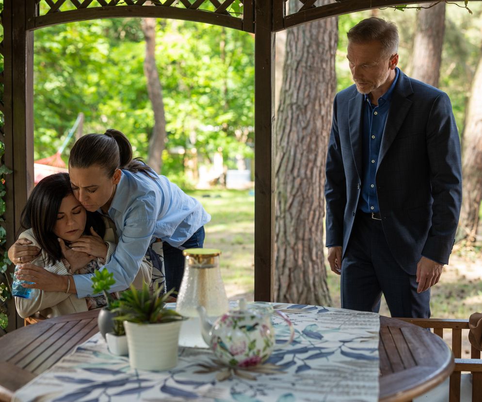 Kinga Zduńska i Magda Budzyńska w emocjonalnej scenie wsparcia, siedzące przy stole w ogrodowej altanie, z Andrzejem Budzyńskim stojącym w tle. Kinga jest wyraźnie poruszona informacjami dotyczącymi Hoffmana. Opis odcinka i inne newsy dostępne na Super Seriale.