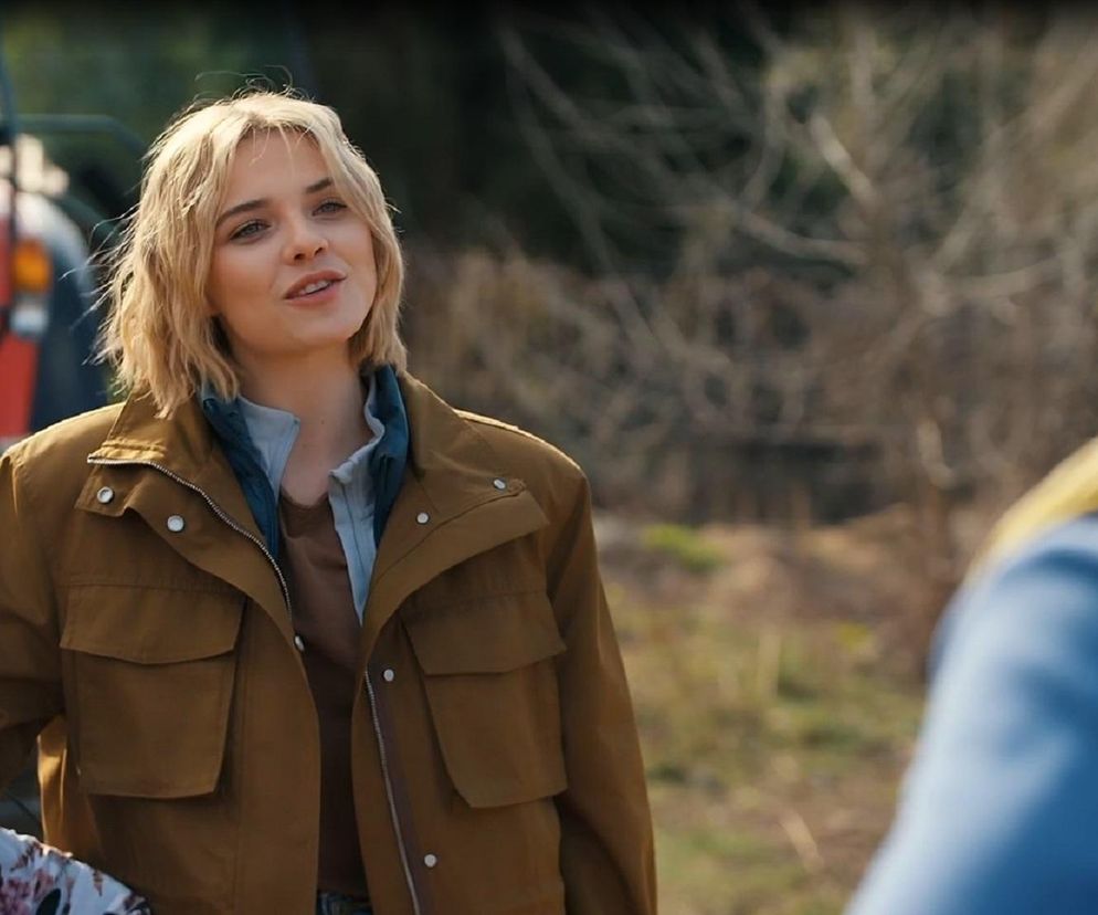 Oliwia Drożdżyk jako Janka, młoda blondynka w brązowej kurtce, stoi na tle traktora, rozmawiając z mężczyzną widocznym z tyłu. Aktorka w scenie z serialu Barwy szczęścia, której fabułę można śledzić na Super Seriale.