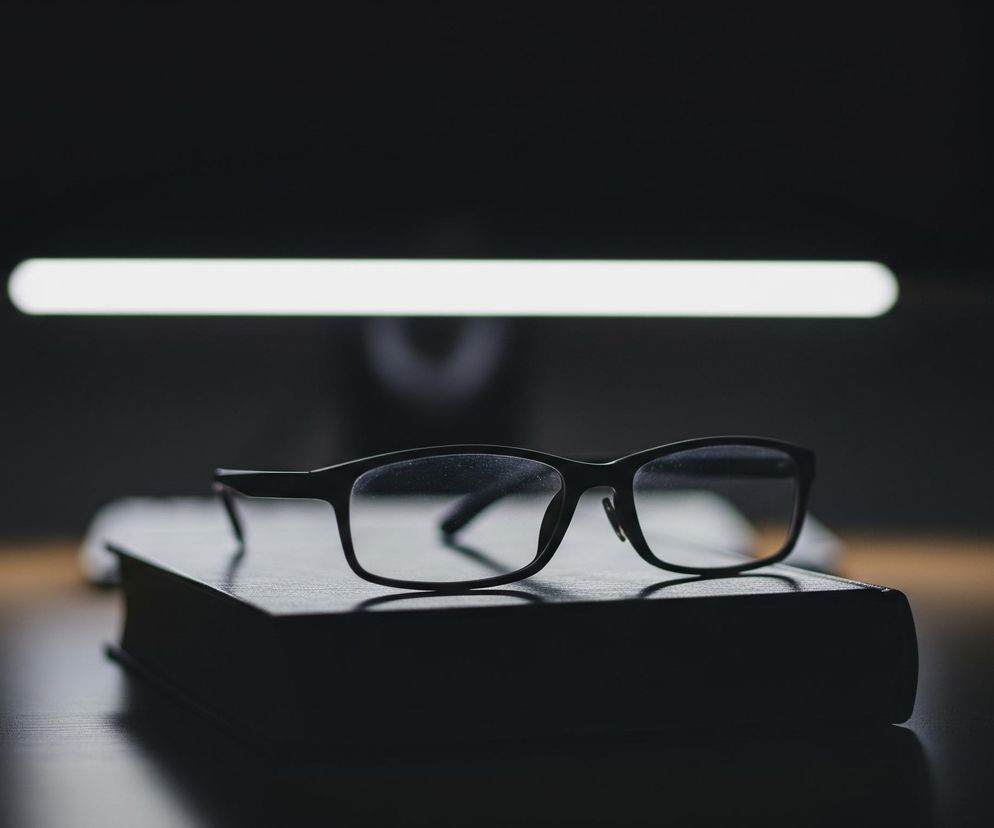 Czarne okulary leżą na ciemnej książce, która spoczywa na drewnianym blacie. Tło jest ciemne, z rozmyciem, a tuż nad okularami widoczna jest pozioma, jasna linia światła, prawdopodobnie z monitora lub lampki biurkowej. Blat jest delikatnie podświetlony od lewej strony, tworząc ciepły, brązowawy odcień. Całość utrzymana jest w ciemnej tonacji, podkreślającej kontrast między jasnym światłem a otaczającą ciemnością.