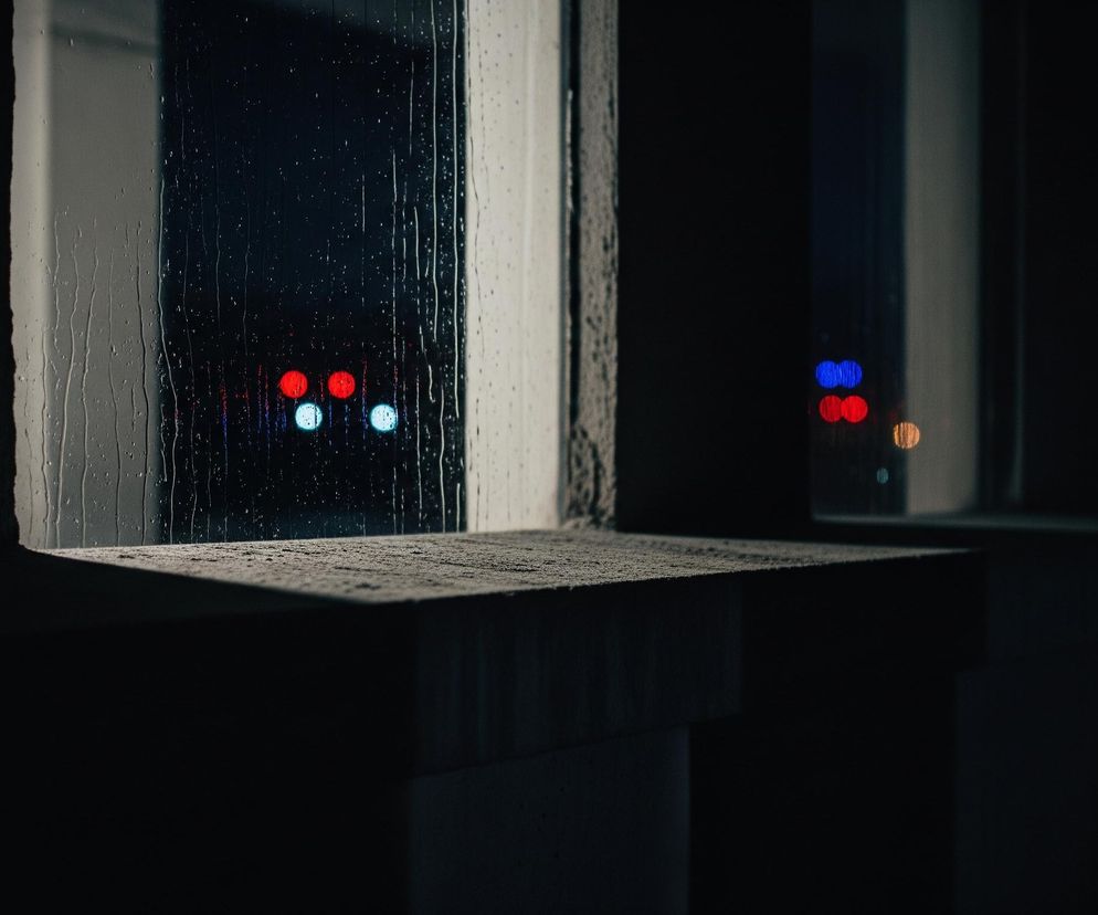 Okno, pokryte kroplami deszczu, zajmuje centralną część obrazu, ukazując rozmyte światła w tle. Po lewej stronie szyby widoczne są dwa czerwone i dwa niebieskie, okrągłe światła, natomiast po prawej, przez drugą szybę, widać dwa niebieskie i dwa czerwone światła oraz jedno żółtawe. Dolna część obrazu przedstawia betonowy parapet lub krawędź ściany, częściowo oświetlony, co nadaje mu teksturę i cienie. Cała scena jest utrzymana w ciemnych barwach, z dominującą czernią i szarościami, podkreślającymi nocny lub deszczowy nastrój.
