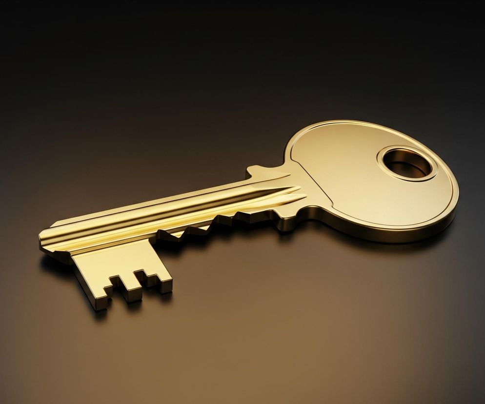 Złoty klucz leży na ciemnym, błyszczącym tle, dominując kompozycję. Jego powierzchnia odbija światło, tworząc jasne refleksy wzdłuż trzonu i na okrągłej główce z otworem. Cień klucza jest widoczny na tle, pogłębiając wrażenie trójwymiarowości.