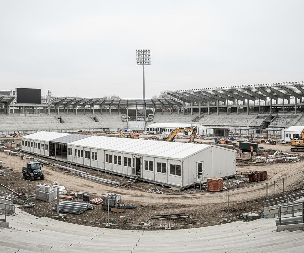 Rozległy stadion sportowy z widownią zbudowaną z betonowych stopni otacza owalny plac, który jest obecnie placem budowy. Na placu budowy znajdują się tymczasowe białe pawilony, w tym długi modułowy budynek w centrum i kilka mniejszych po bokach, oraz ciężki sprzęt budowlany, taki jak żółte koparki i traktor, a także stosy materiałów budowlanych. Nad stadionem wznosi się wysoki maszt oświetleniowy z licznymi reflektorami, a po lewej stronie widoczny jest duży, czarny ekran. Całość znajduje się pod jasnym, zachmurzonym niebem.