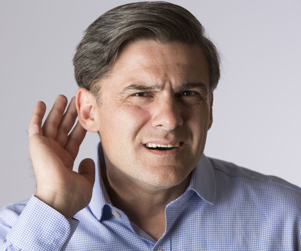 Mężczyzna w średnim wieku z siwymi włosami i marsową miną, trzymający dłoń przy uchu, symbolizuje problemy ze słuchem i niedosłuch u dorosłych. Zdjęcie ilustruje wyzwania związane z percepcją dźwięku, o których można przeczytać na portalu Poradnik Zdrowie.