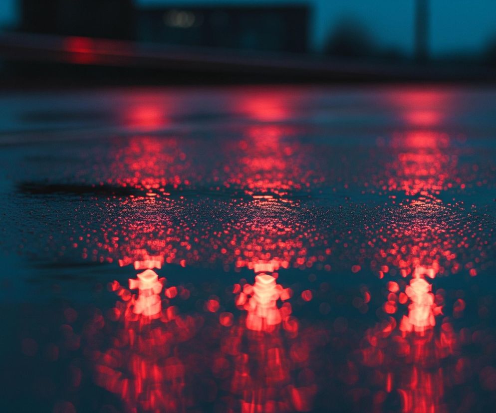 Mokra, ciemna powierzchnia odbija intensywne, czerwone światła, które tworzą nieregularne, rozmyte plamy i smugi na pierwszym planie oraz w oddali. Liczne drobne krople wody pokrywają całą widoczną płaszczyznę, rozpraszając światło na małe punkty. W tle, powyżej horyzontu wyznaczonego przez odbicia, widoczne są zamglone, ciemne kształty, sugerujące budynki lub inne obiekty, zanurzone w niebieskawo-granatowej poświacie.