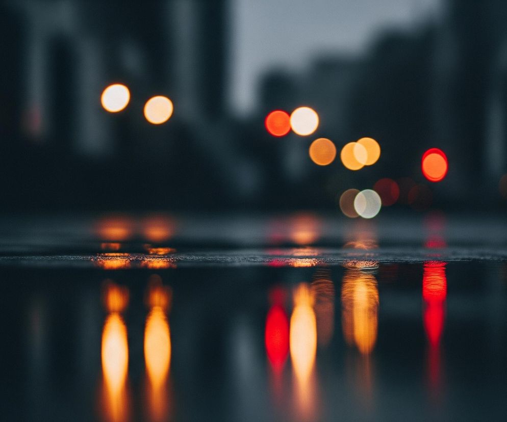 Na pierwszym planie widoczna jest mokra powierzchnia, prawdopodobnie asfalt, odbijająca światła w oddali. Powierzchnia ta jest ciemnoszara, a jej faktura sugeruje obecność kropli wody lub kałuż. Nad nią, w tle, znajduje się rozmyty obraz miejskiego krajobrazu, z niewyraźnymi konturami budynków lub drzew.

Główne obiekty to liczne, rozmyte plamy światła o różnych kolorach: pomarańczowych, białych i czerwonych, tworzące efekt bokeh. Te światła są odbite w mokrej nawierzchni na dole kadru, co tworzy wydłużone, pionowe smugi świetlne. Cały obraz utrzymany jest w ciemnej, wieczorowej lub nocnej tonacji.