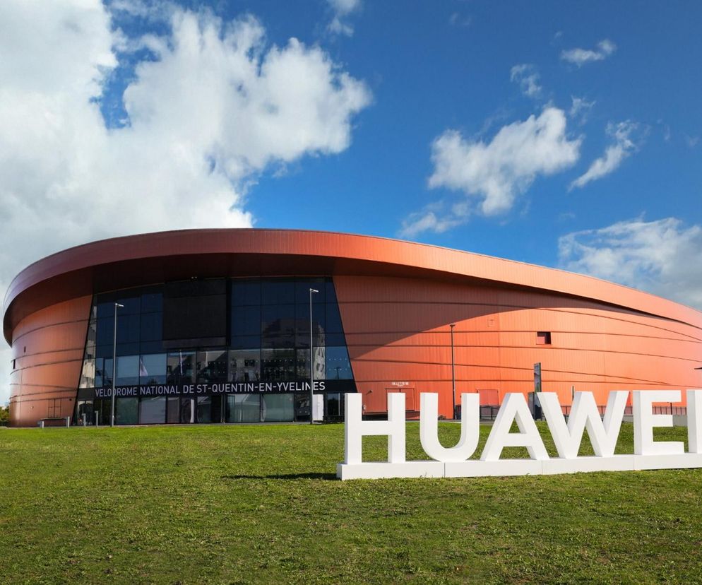 Pomarańczowo-brązowy, owalny budynek z dużymi, przeszklonymi drzwiami wejściowymi, przed którym na trawniku widnieje duży biały napis HUAWEI. Obiekt widoczny jest w tle, a na pierwszym planie znajduje się trawa. Informacje o nowych smartwatchach Huawei przeczytasz na Super Biznes.