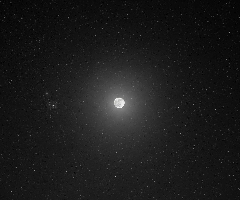 Pełnia księżyca dominuje centralną część obrazu, emanując jasnym, białym światłem i otoczona delikatną poświatą. Całe tło stanowi ciemne, gwieździste niebo, usiane niezliczonymi, drobnymi, białymi punktami, symbolizującymi gwiazdy. W lewej, środkowej części widoczne jest niewielkie zgrupowanie jaśniejszych gwiazd, tworzących delikatną gromadę. Kolorystyka obrazu ogranicza się do bieli, szarości i czerni, tworząc kontrastową scenę kosmiczną.