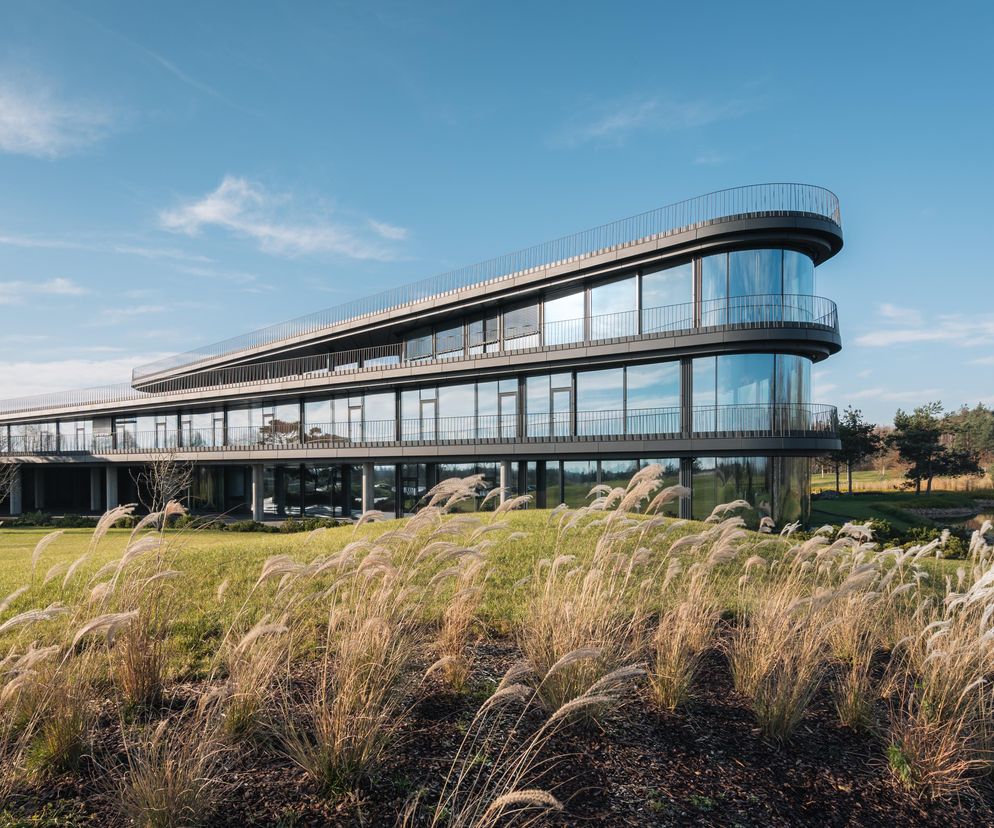 Nowoczesna siedziba Press Glass w Konopiskach, projektu KONIOR STUDIO (Tomasz Konior, Dominik Koroś). Horyzontalna bryła z przeszkloną elewacją, otoczona trawami ozdobnymi i zielonym terenem, harmonijnie wpisuje się w krajobraz pola golfowego, o czym przeczytasz na Architektura Murator Plus.