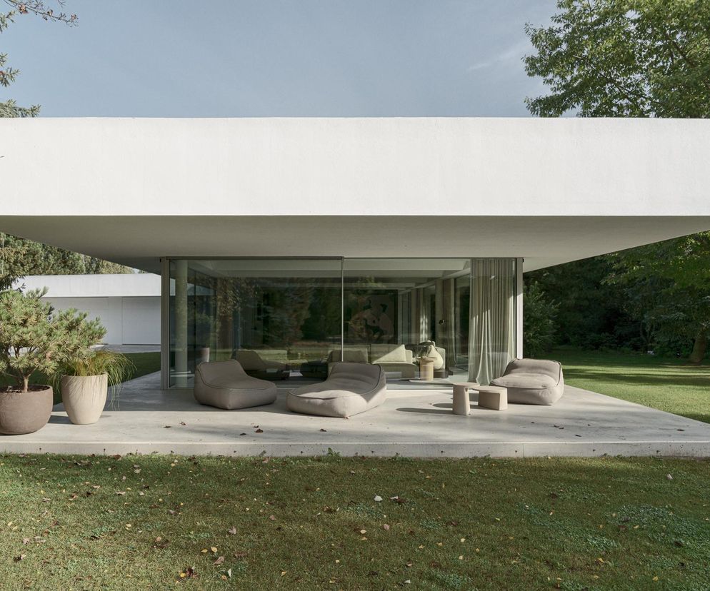 Minimalistyczny Dom S w kształcie litery S, zrealizowany przez PL.ARCHITEKCI, z białym, horyzontalnym dachem i przeszklonymi ścianami. Przed budynkiem betonowy taras z nowoczesnymi meblami wypoczynkowymi, otoczony zielonym ogrodem. Więcej o architekturze tego typu można przeczytać na Architektura Murator Plus.