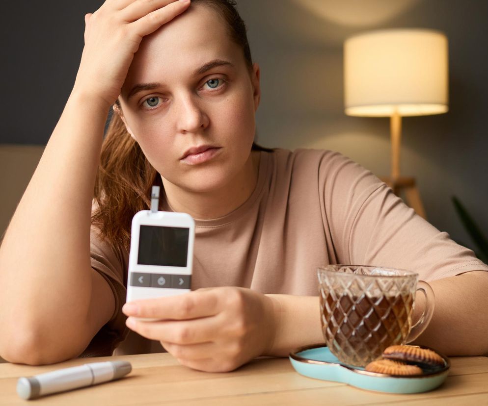 Kobieta z cukrzycą typu 1 siedzi przy stole, trzymając w ręku glukometr i patrząc wprost na widza. Obok leżą nakłuwacz oraz filiżanka z herbatą i ciastkami. Zdjęcie symbolizuje trudności pacjentów z cukrzycą w walce o refundację pomp insulinowych, o czym więcej przeczytasz na Poradnik Zdrowie.
