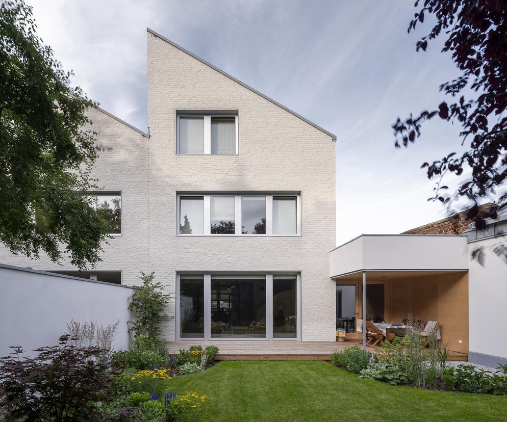 Elewacja bliźniaka w Wilanowie autorstwa Jojko+Nawrocki Architekci, z jasnych płytek silikatowych i skośnym dachem, harmonizuje z otoczeniem. Przed domem zielony trawnik i taras, a obok drzewa. Więcej o architekturze na portalu Architektura Murator Plus.