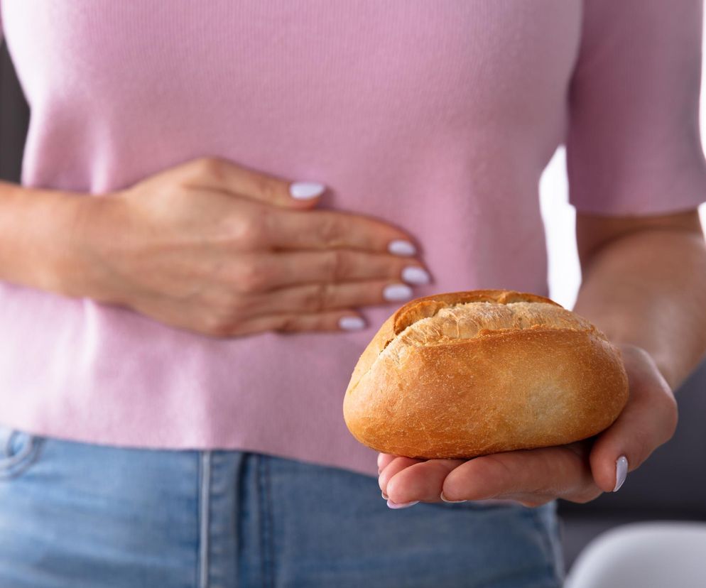 Kobieta w różowej bluzce trzymająca świeżą bułkę w jednej dłoni i drugą dłoń na brzuchu, symbolizując ból brzucha po zjedzeniu pieczywa. Ta ilustracja odnosi się do problemu nadwrażliwości na zboża, o której więcej przeczytasz na Poradnik Zdrowie.