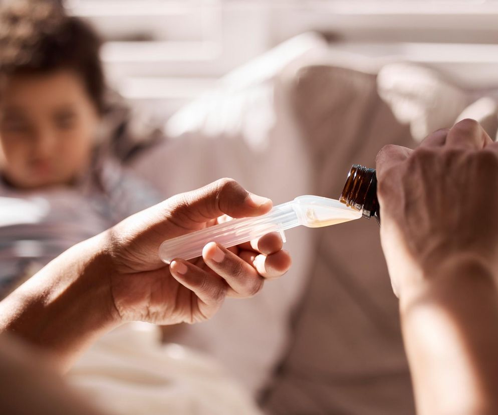 Osoba dorosła nalewająca syrop do miarki, z niewyraźną postacią dziecka w tle. Scena podkreśla problem bezpieczeństwa leków i ich podawania, nawiązując do artykułu o skażonym syropie, co można przeczytać na portalu Poradnik Zdrowie.