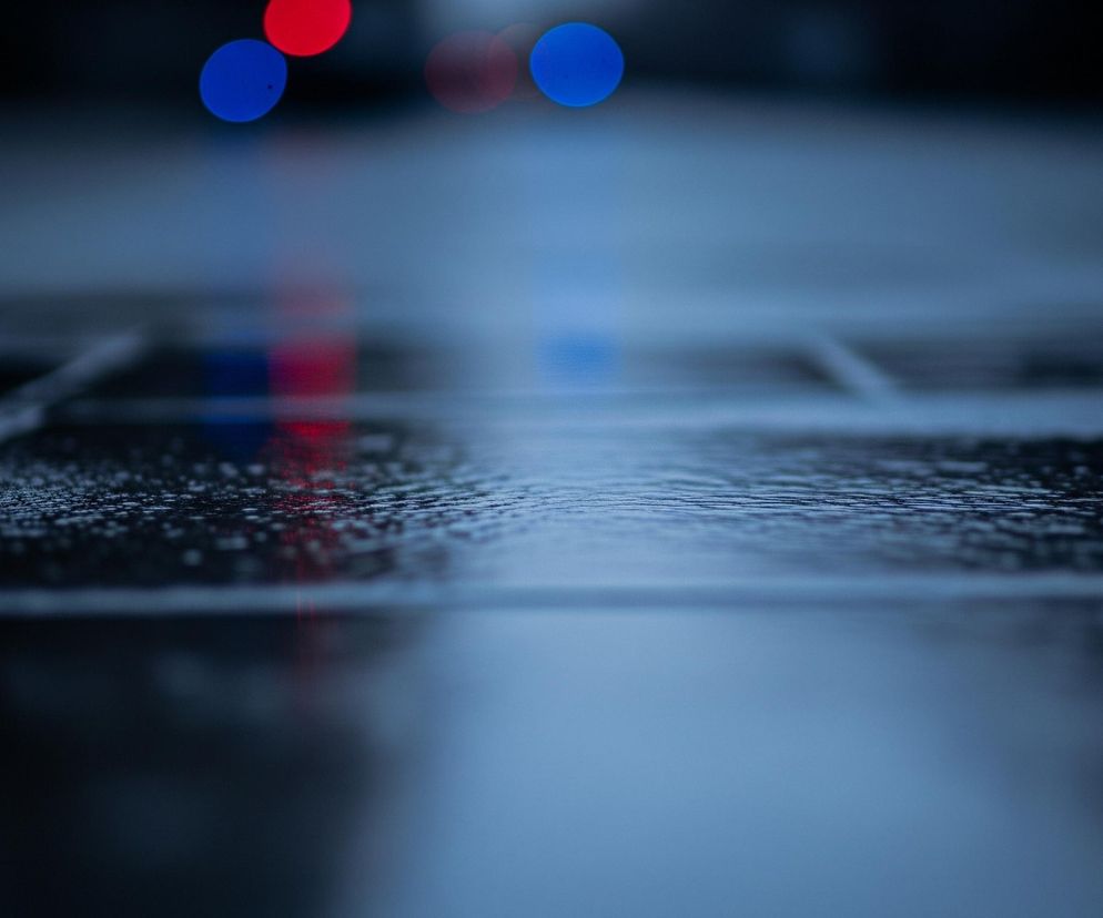 Ulica pokryta deszczem lub kałużami, z widocznymi odbiciami światła. Powierzchnia drogi, złożona z prostokątnych płyt, jest mokra i błyszcząca, z wyraźnie widocznymi kroplami wody w górnej części kadru. Dalej, w tle, pojawiają się rozmyte, kuliste światła: dwa czerwone i dwa niebieskie, które odbijają się w wodzie na nawierzchni, tworząc podłużne smugi. Dominują ciemne odcienie błękitu i granatu, z akcentami jaskrawej czerwieni i błękitu świateł.