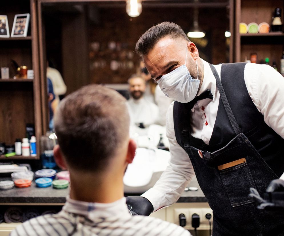 Fryzjer w białej koszuli, czarnej kamizelce, dżinsowym fartuchu i masce ochronnej z czarnymi rękawiczkami przygotowuje się do strzyżenia klienta, który siedzi tyłem do kamery. Scena z wnętrza salonu fryzjerskiego, wspierająca akcję #ChceDoSalonu. Więcej o akcji przeczytasz na portalu Poradnik Zdrowie.