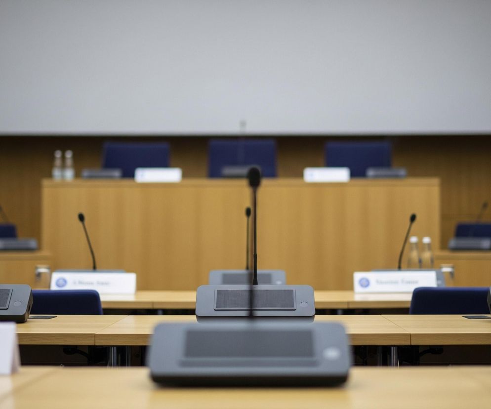 Rząd rzędem drewnianych stołów konferencyjnych z mikrofonami i tabliczkami z nazwiskami rozciąga się w poprzek kadru. Na każdym stole widoczne są szare pulpity z wbudowanymi głośnikami i giętkimi mikrofonami na długich ramionach, a także szklane butelki wody i puste szklanki. Za stołami stoją rzędy krzeseł z ciemnoniebieskim obiciem, a w tle znajduje się duży, jasny ekran projekcyjny umieszczony nad drewnianym panelem ściennym.