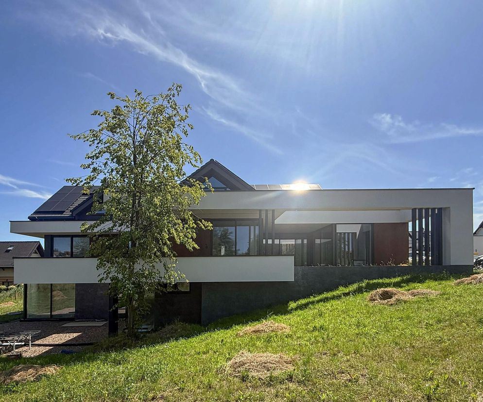 Nowoczesny, jednorodzinny budynek mieszkalny w Modlnicy projektu KJ Architekci, z płaskim dachem ukrytym za białymi ramami, dużymi oknami i panelami fotowoltaicznymi. Otoczony zielonym terenem, prezentuje architekturę, o której można przeczytać na Architektura Murator Plus.