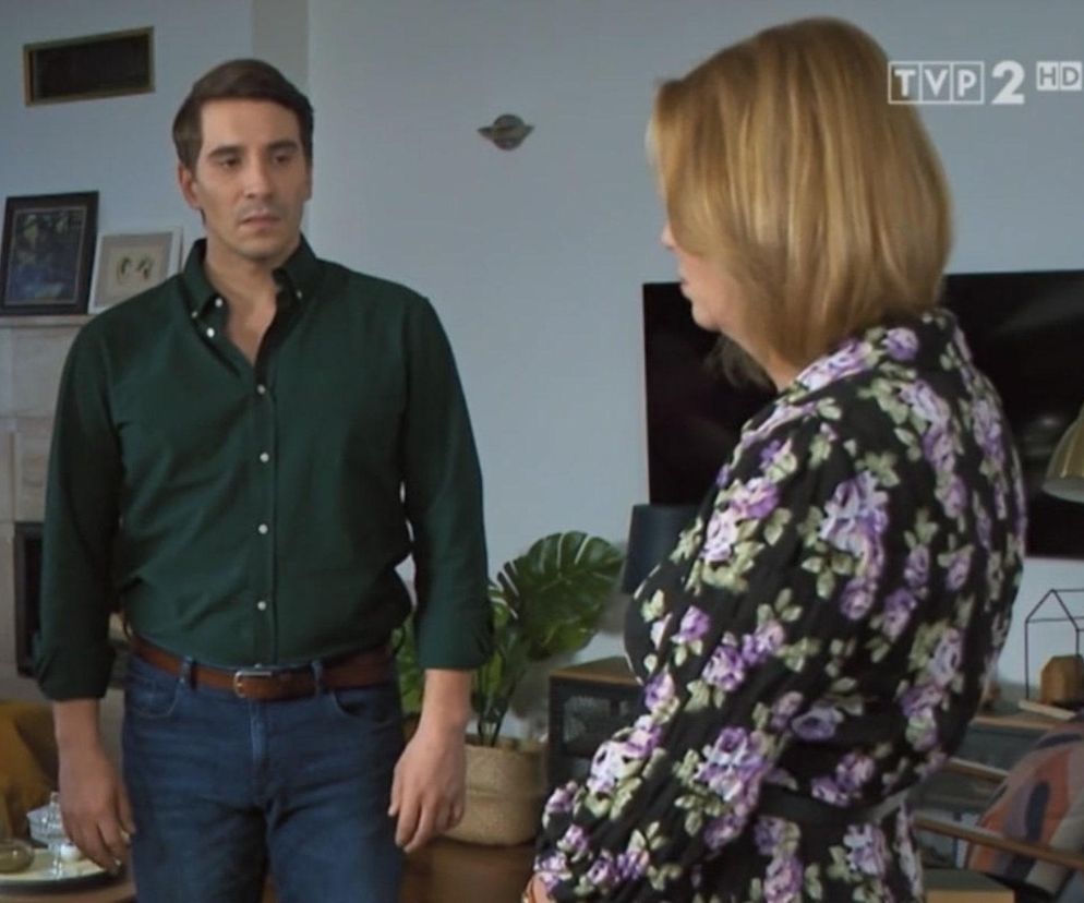 Tomasz Kępski, ubrany w zieloną koszulę i jeansy, stoi w mieszkaniu obok Wandy, która ma na sobie kwiecistą sukienkę. Scena z serialu Barwy szczęścia przedstawia moment konfliktu, gdzie Kępski staje w obliczu oburzonej matki, wyrażając napięcie i trudne emocje. Więcej o ich losach przeczytasz na Super Seriale.