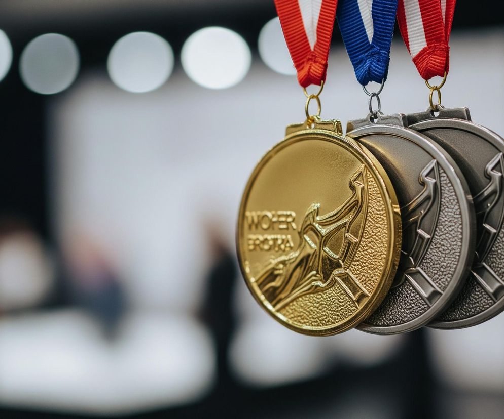 Trzy medale wiszą obok siebie na barwnych wstążkach. Po lewej stronie znajduje się złoty medal z wytłoczonym napisem WOHER EROBA i stylizowanym wzorem. Obok niego wisi srebrny medal, a następnie brązowy medal, oba z podobnymi reliefami i fakturą. Medale zawieszone są na wstążkach w kolorach czerwonym, białym i niebieskim, widocznych u góry kadru, na tle rozmytego, jasnego otoczenia z okrągłymi światłami bokeh.
