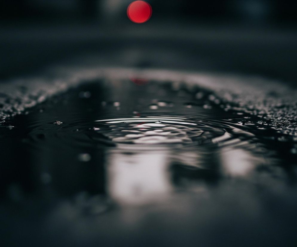 Centralnym elementem jest kałuża deszczowej wody na ciemnej, szorstkiej nawierzchni, prawdopodobnie asfalcie, rozciągająca się od dołu obrazu. Na powierzchni wody widoczne są koncentryczne fale, świadczące o niedawno spadłej kropli lub innym zakłóceniu. W oddali, wysoko na ciemnym tle, unosi się wyraźna, czerwona plama światła, a pod nią, niewyraźne odbicie tego światła jest widoczne na powierzchni kałuży. Całość ma ciemną, stonowaną kolorystykę z dominującymi odcieniami szarości i czerni, przełamaną jaskrawą czerwienią.