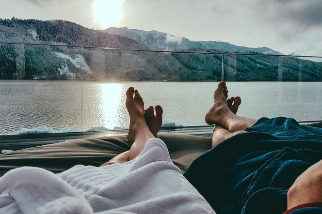 Stopy pary w białym i niebieskim szlafroku leżące na leżakach z widokiem na jezioro i zaśnieżone góry o zachodzie słońca. Obraz symbolizuje relaks i wakacyjny wyjazd, a o formalnościach przed podróżą przeczytasz na Poradnik Zdrowie.