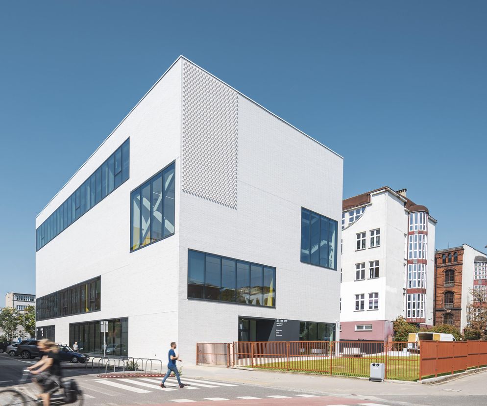 Nowoczesny, prostopadłościenny budynek w białej cegle arch_it Piotra Zybury, z dużymi oknami, obok historycznej zabudowy we Wrocławiu. To hybrydowy zespół sal gimnastycznych przy XIII Liceum, o którym można przeczytać na Architektura Murator Plus.