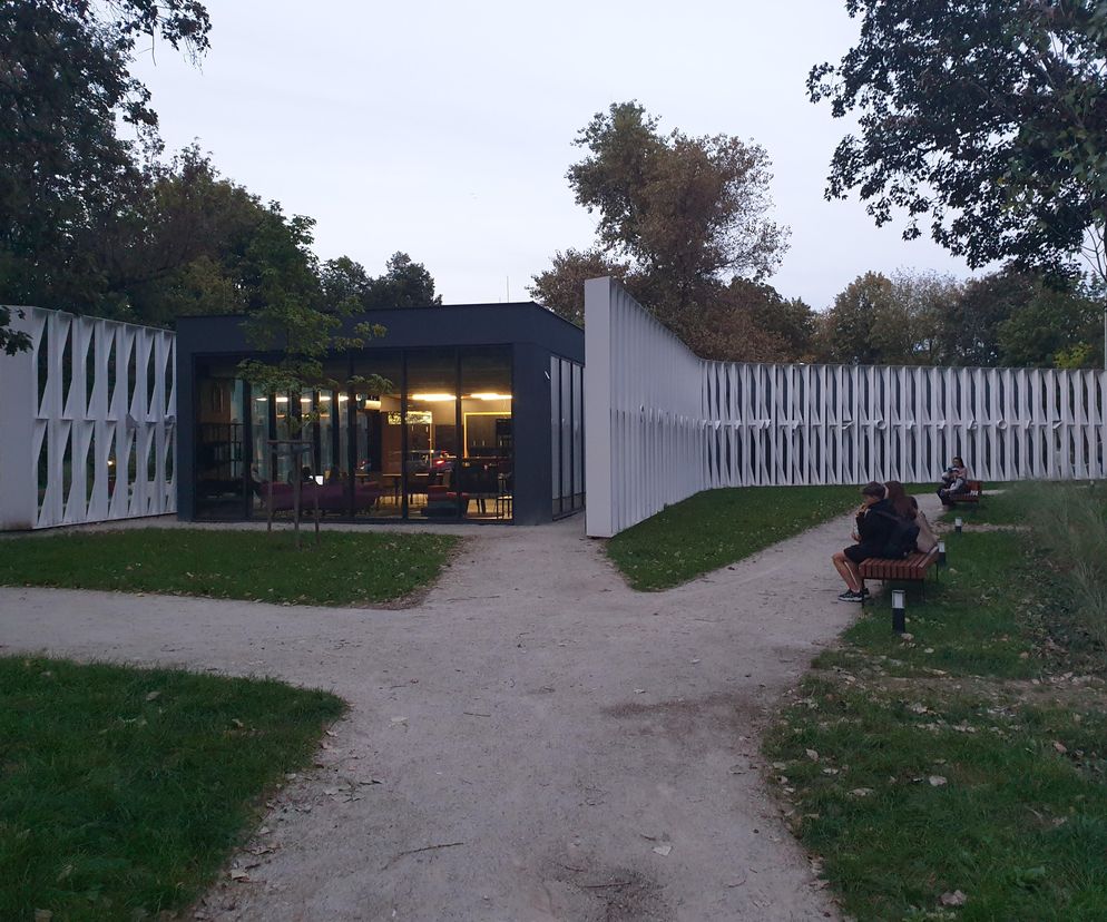 Centrum Lokalne Żoliborz, widoczne o zmierzchu, z minimalistyczną elewacją i przeszkleniami, które rozświetlają wnętrze. Białe, ażurowe ściany parawanowe filtrują światło. Ludzie odpoczywają na ławkach na pierwszym planie. Więcej o architekturze tego obiektu znajdziesz na Architektura Murator Plus.