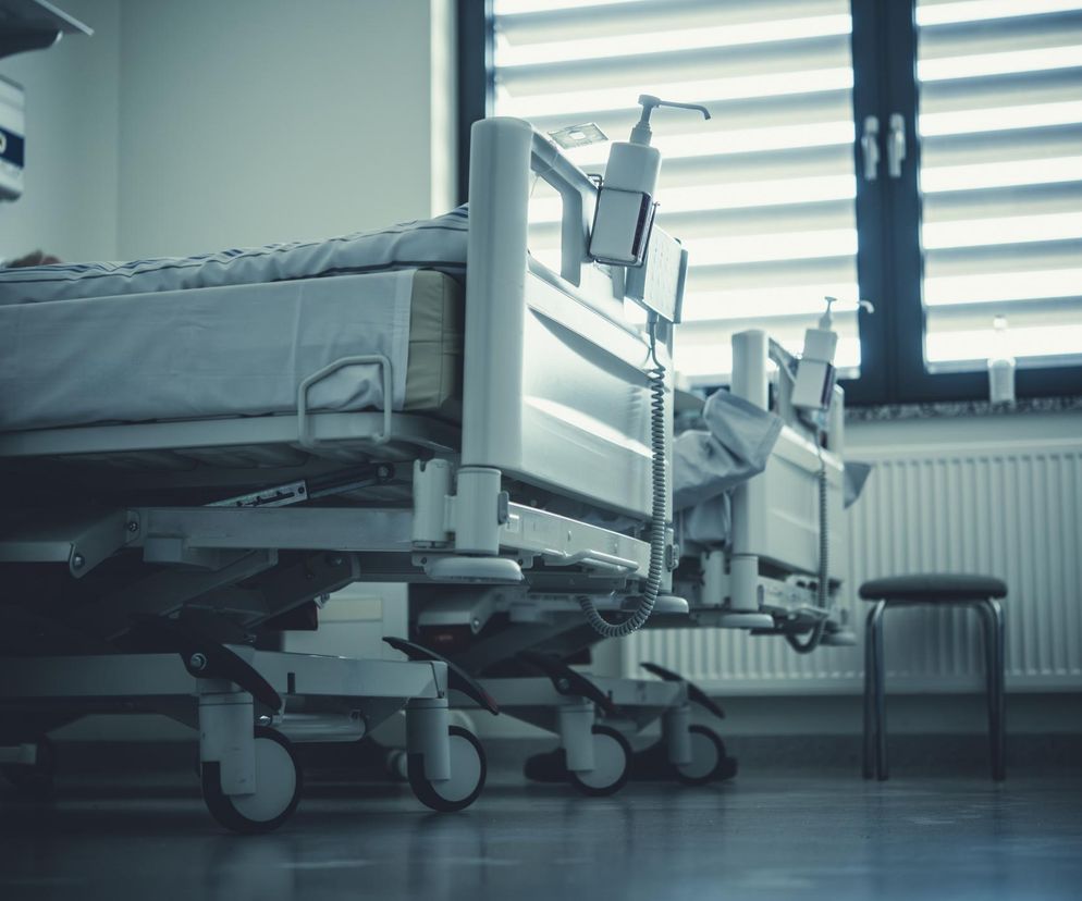 Puste łóżka szpitalne w sali pacjenta, symbolizujące wagę rzetelnej dokumentacji medycznej w kontekście zatrzymania ordynatora szpitala wojewódzkiego. W tle widoczne okno z żaluzjami i grzejnik. Więcej o tej sprawie przeczytasz na portalu Poradnik Zdrowie.