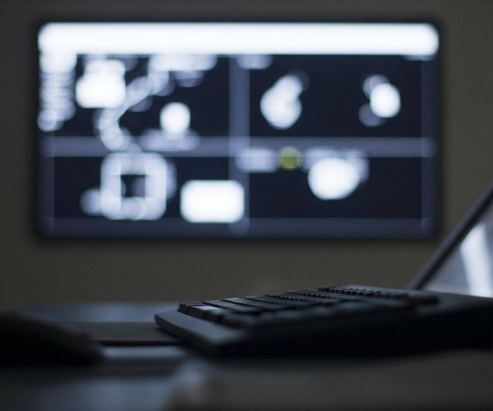 Centralnym elementem zdjęcia jest rozmazany, podświetlony ekran widoczny w tle, prezentujący abstrakcyjne białe kształty na ciemnym tle, przypominające schemat lub interfejs użytkownika. Na pierwszym planie, po prawej stronie, ostro zarysowana jest ciemna klawiatura komputera z widocznymi rzędami klawiszy, a obok niej, po lewej stronie, leży myszka komputerowa. Całość utrzymana jest w ciemnej tonacji, z dominacją szarości i czerni, a światło pochodzi głównie z ekranu w tle.