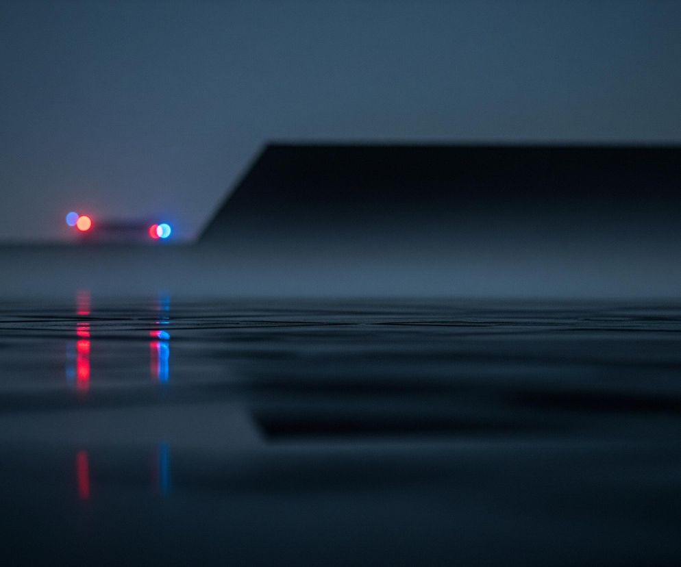 Ciemną, stonowaną scenę z dominującym granatowo-szarym tłem. W oddali, po lewej stronie, widoczne są cztery rozmyte, okrągłe światła: dwa czerwone i dwa niebieskie, ułożone w parach poziomo. Te światła odbijają się w falującej, ciemnej powierzchni na pierwszym planie, tworząc wydłużone, rozmyte smugi czerwieni i błękitu. Po prawej stronie, w tle, z ciemności wyłania się czarny, geometryczny kształt, przypominający krawędź budynku lub konstrukcji, która częściowo przysłania tło. Cała scena jest zdominowana przez ciemne, chłodne odcienie i sugeruje nocne lub bardzo zmierzchowe otoczenie.