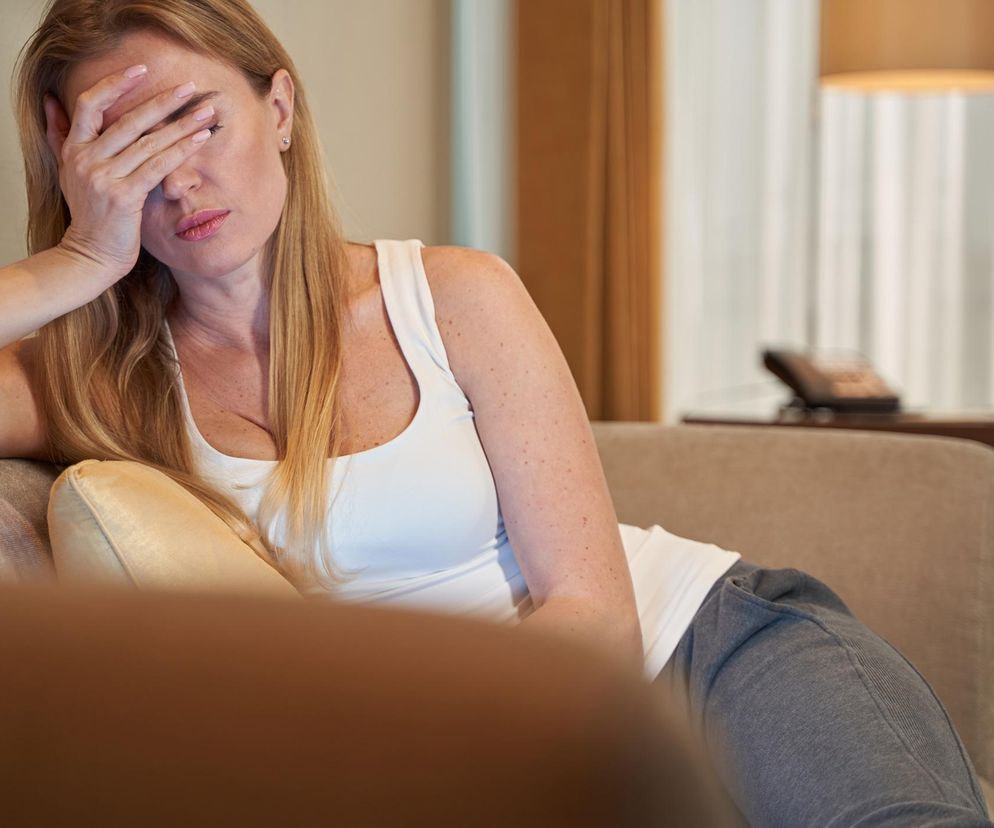Blondwłosa kobieta w białej koszulce i szarych spodniach dresowych leży na beżowej sofie, z dłonią zakrywającą twarz, wyrażając zmęczenie lub ból głowy. Obok niej beżowa poduszka. Scena symbolizuje objawy długiego COVID, takie jak przewlekłe zmęczenie i mgła mózgowa, o których można przeczytać na portalu Poradnik Zdrowie.