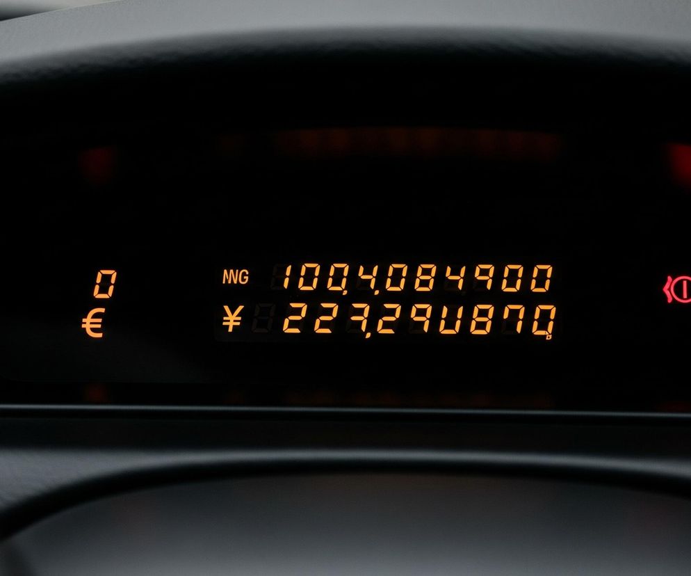 Cyfrowy wyświetlacz z jasnopomarańczowymi znakami jest głównym elementem obrazu, umieszczonym na czarnym tle. Górna linia tekstu pokazuje NNG 100,4084900, a pod nią widnieje symbol japońskiego jena (¥) oraz liczba 227,290870. Po lewej stronie ekranu widać cyfrę 0 i symbol euro (€) poniżej niej. W prawym górnym rogu ekranu świeci się czerwona ikona przypominająca wykrzyknik w kółku, otoczony dwoma półkolami. Całość otacza ciemna, matowa obudowa, z której widoczne są fragmenty gładkich, czarnych powierzchni.