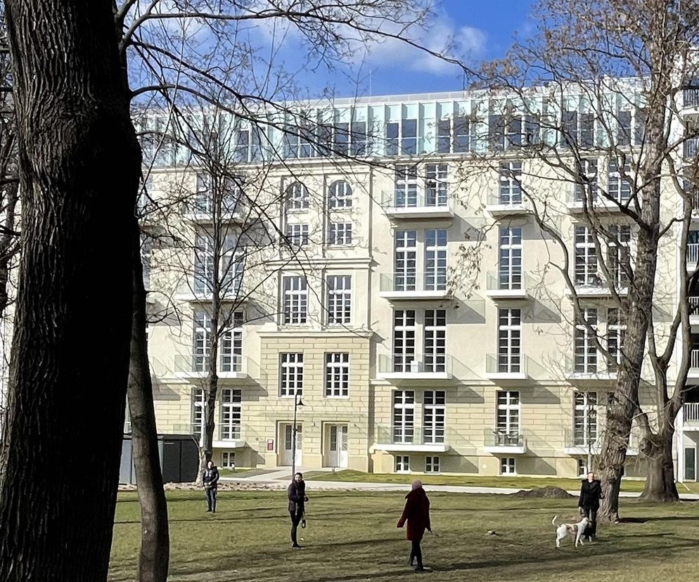 Budynek mieszkalno-usługowy w dawnym kompleksie szpitalnym im. Babińskiego we Wrocławiu, z jasną elewacją i licznymi oknami, otoczony parkiem z drzewami bez liści. Przed budynkiem na trawniku spacerują ludzie z psami, co można przeczytać na Architektura Murator Plus.